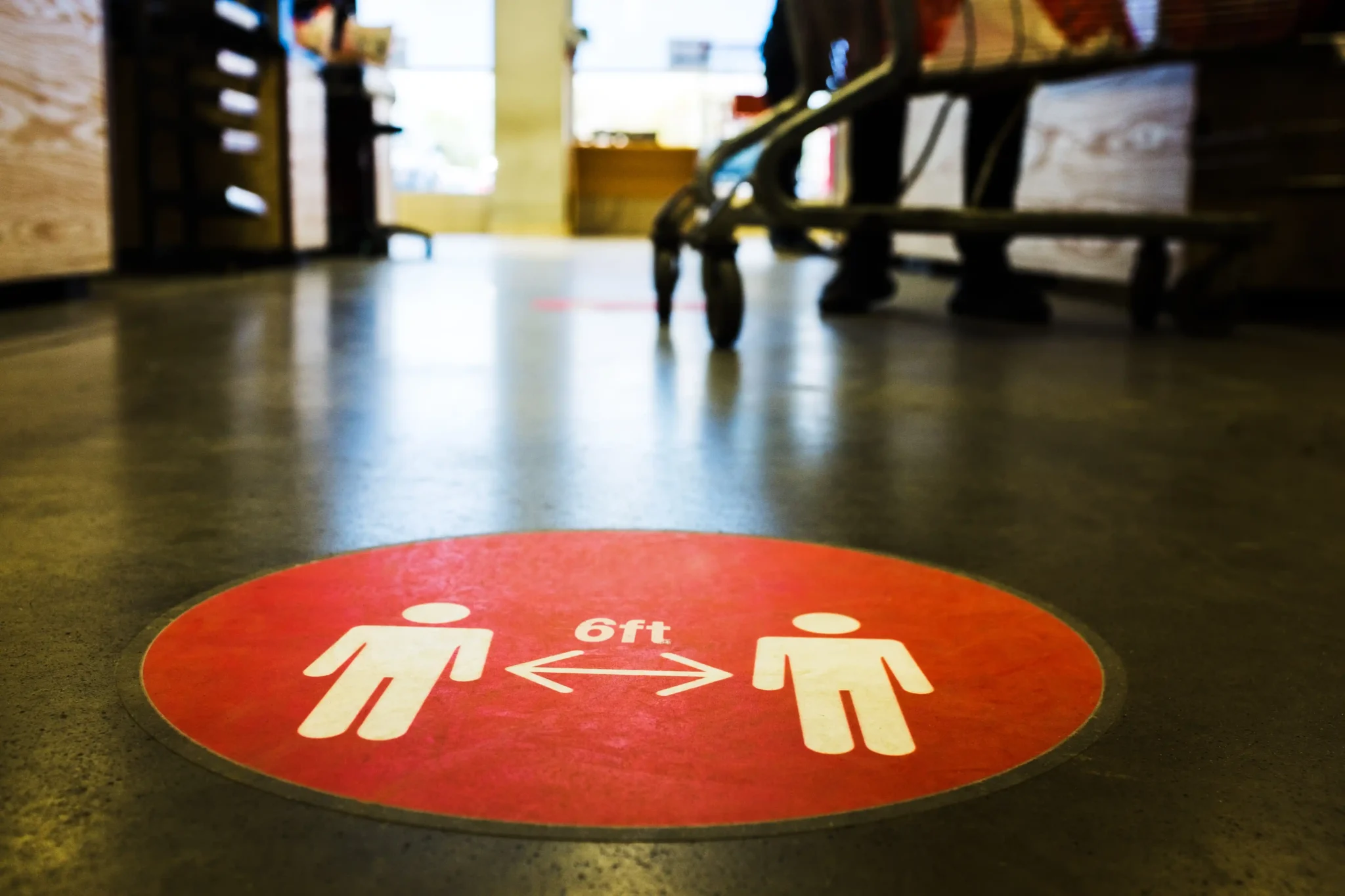 In an Oregon store aisle, a red floor sign with white figures and an arrow highlights the 6-foot social distancing guideline. A shopping cart and person appear in the background, standing on a sturdy concrete coating.