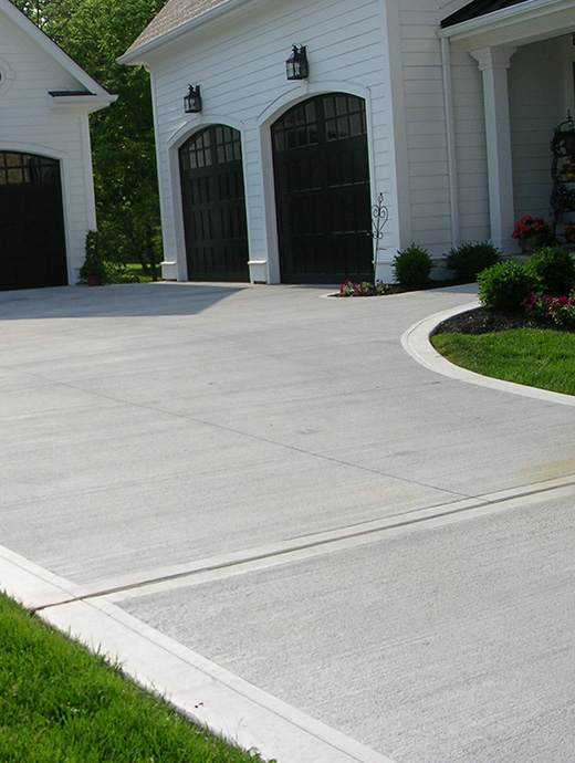 A pristine concrete driveway enhanced with a sleek coating leads to two white garages with dark doors, nestled amidst a lush green lawn and small shrubs, creating an inviting Oregon charm.
