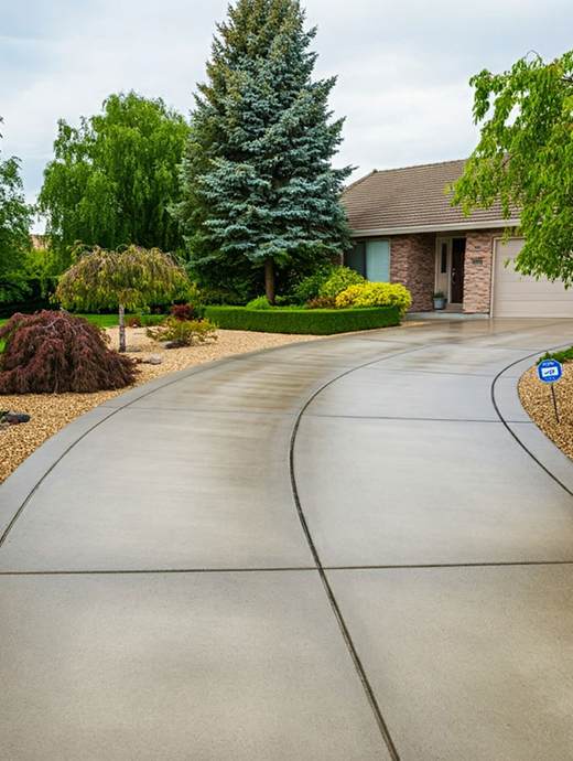 A curving concrete driveway, enhanced with premium concrete coating, leads to a brick house with a garage. It's surrounded by manicured landscaping and towering trees, creating a picturesque scene under Oregon's cloudy sky.
